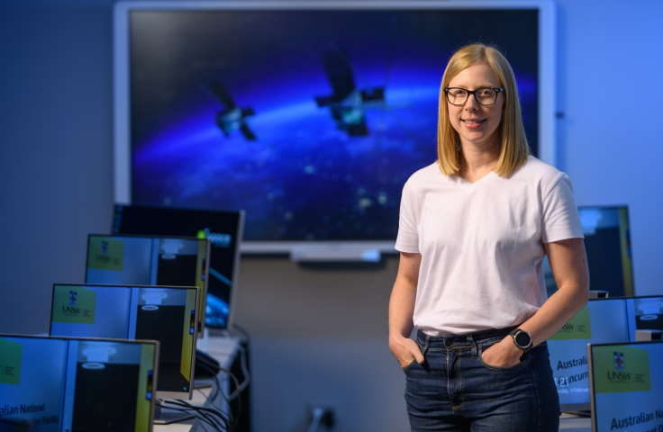 Dr Courtney Bright stands inside a computer lab, smiling with her hands casually in her pockets. Behind her is a large screen featuring a vision of the M2 satellite.